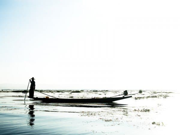 Silhouetted fisherman on a flat, narrow boat on a lake. Man is at the back of the boat, holding a pole in both hands. 