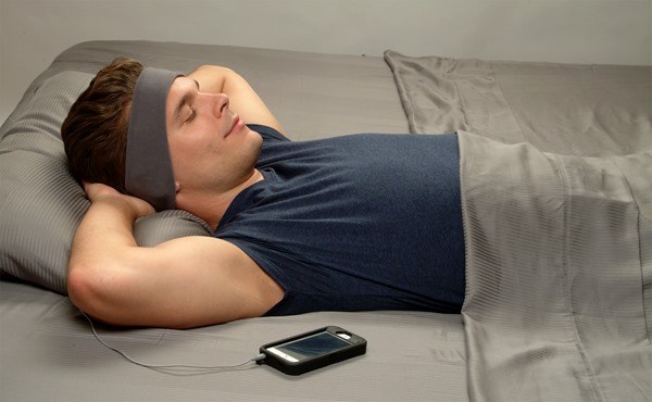 Man lying in bed with hands behind his head, wearing a Sleepphones headband and listening to music on his phone.