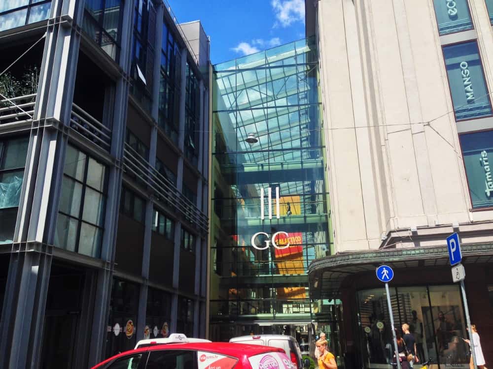 Outside view of Riga Galleria mall, a multistory steel and glass structure. Cars and pedestrians visible in foreground.