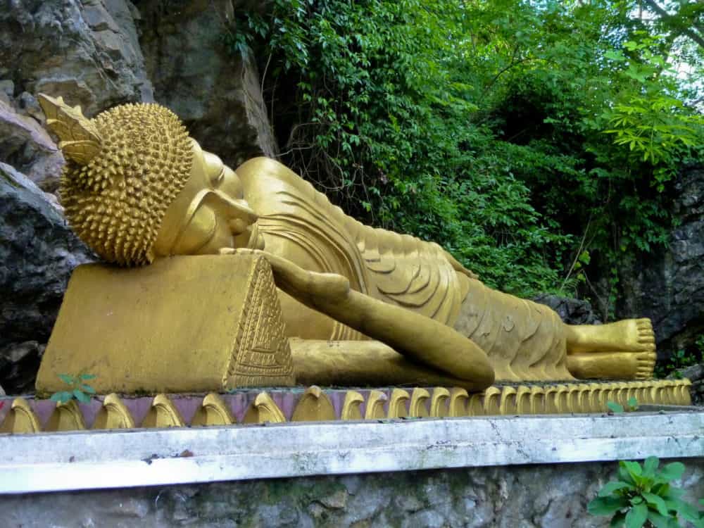 Reclining Buddha statue, covered in gold leaf on a platform near a rocky cliff wall.