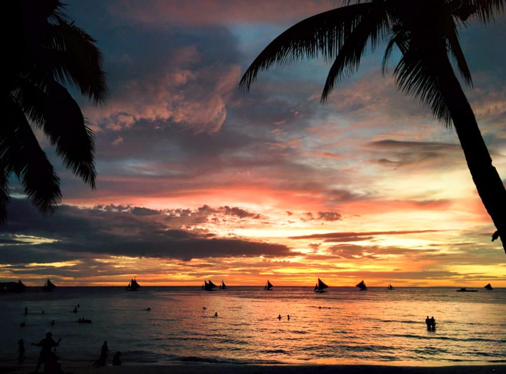 Sunset on a beach in the Philippines, with many small sailboats silhouetted on the horizon and people swimming in the shallows. Palm trees visible at edges of picture.