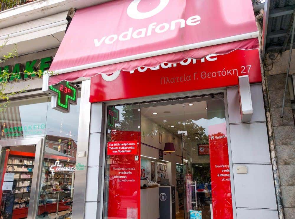 The outside of a small store in Corfu, Greece, with Vodafone branding on the outside. A pharmacy is alongside.