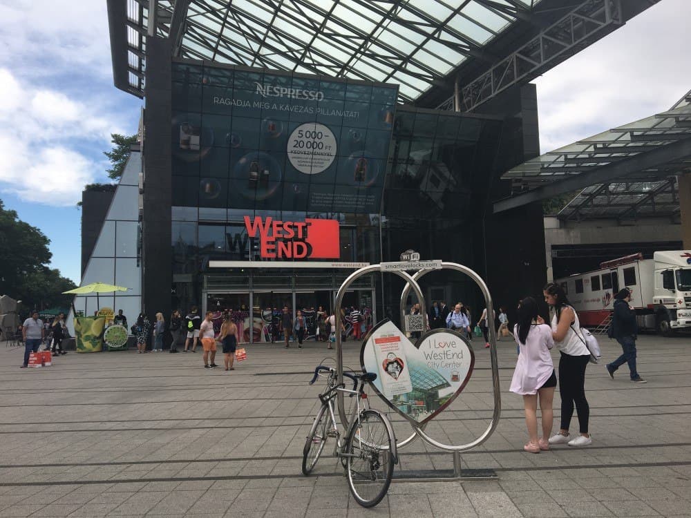 Outside view of the entrance of Westend mall in Budapest, with a large open space outside. Many people are walking in and out of the mall.