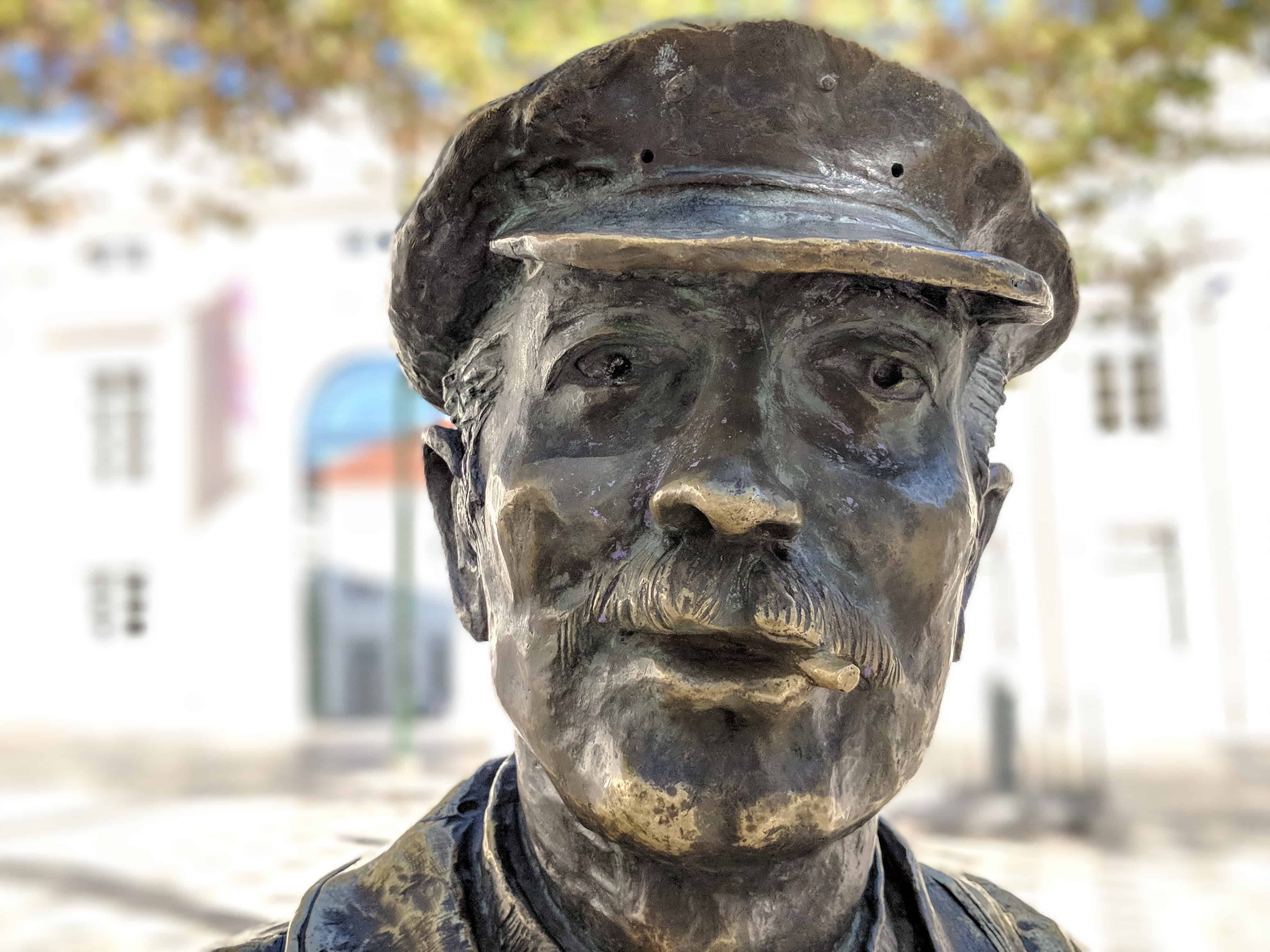 Close-up of the head of a metal statue of a man with a moustache and cap, with blurred background.. 