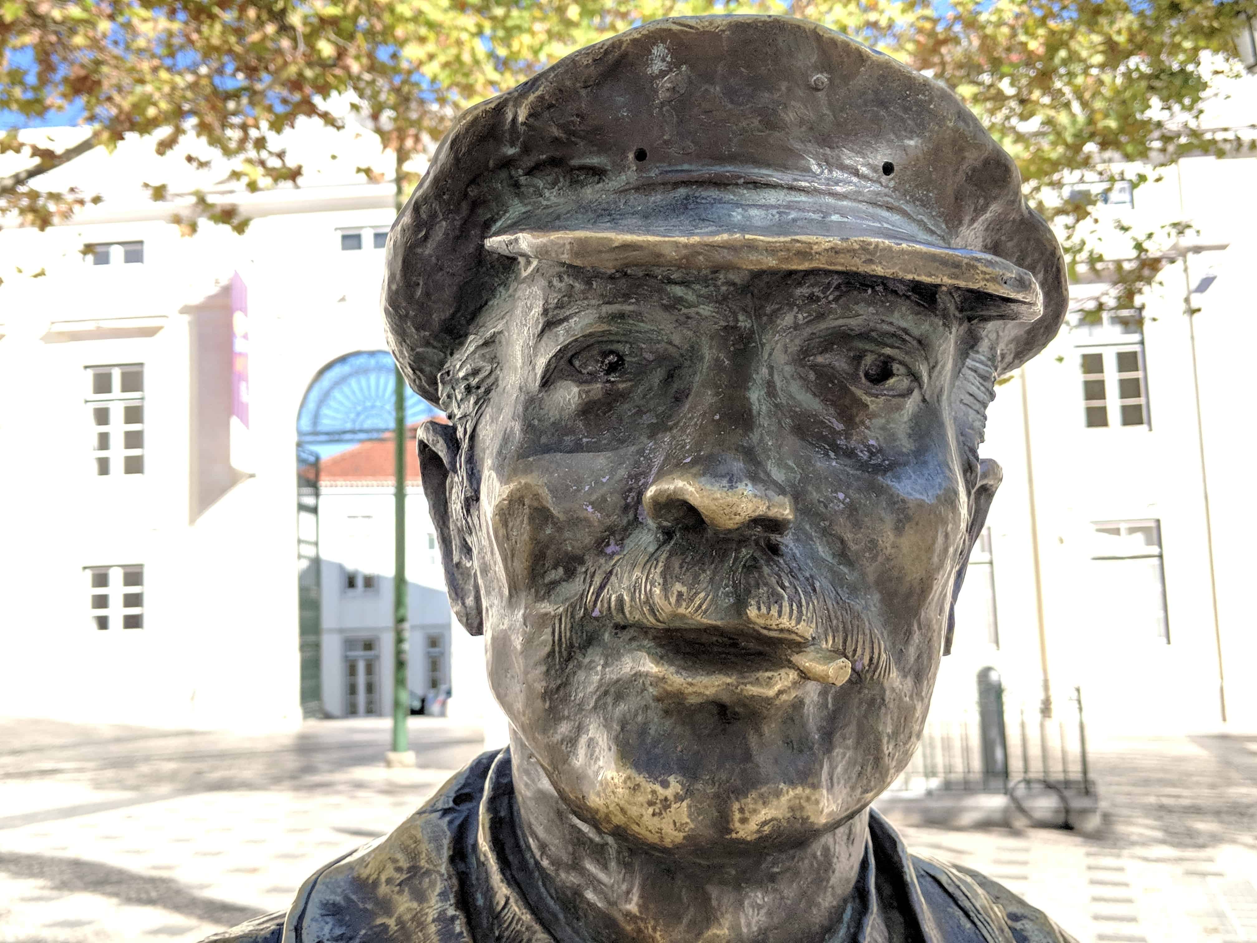 Close-up of the head of a metal statue of a man with a moustache and cap, with white buildings visible behind. 