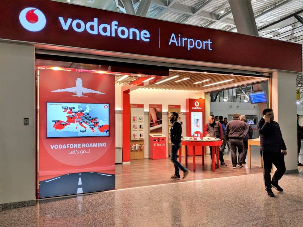 Outside of a store at the airport in Tirana, Albania, with vodafone | Airport in large letters above the entrance. People are visible inside and outside the store.