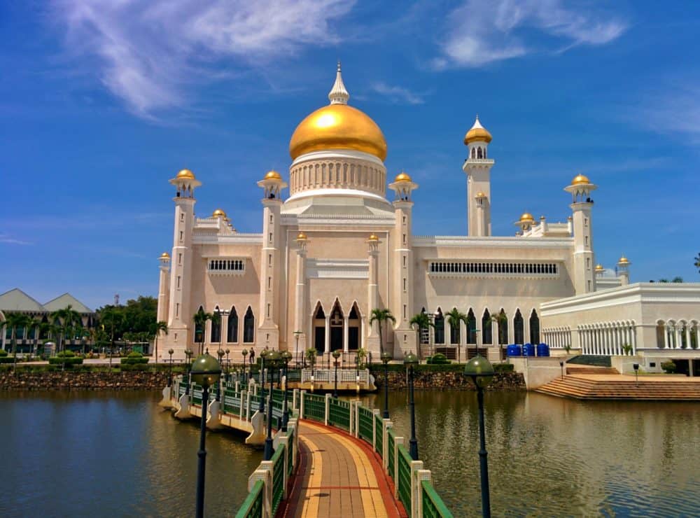 Large white mosque with gold covering at the top of each minaret. A curving footbridge leads over a lake surrounding the mosque. Blue skies behind.