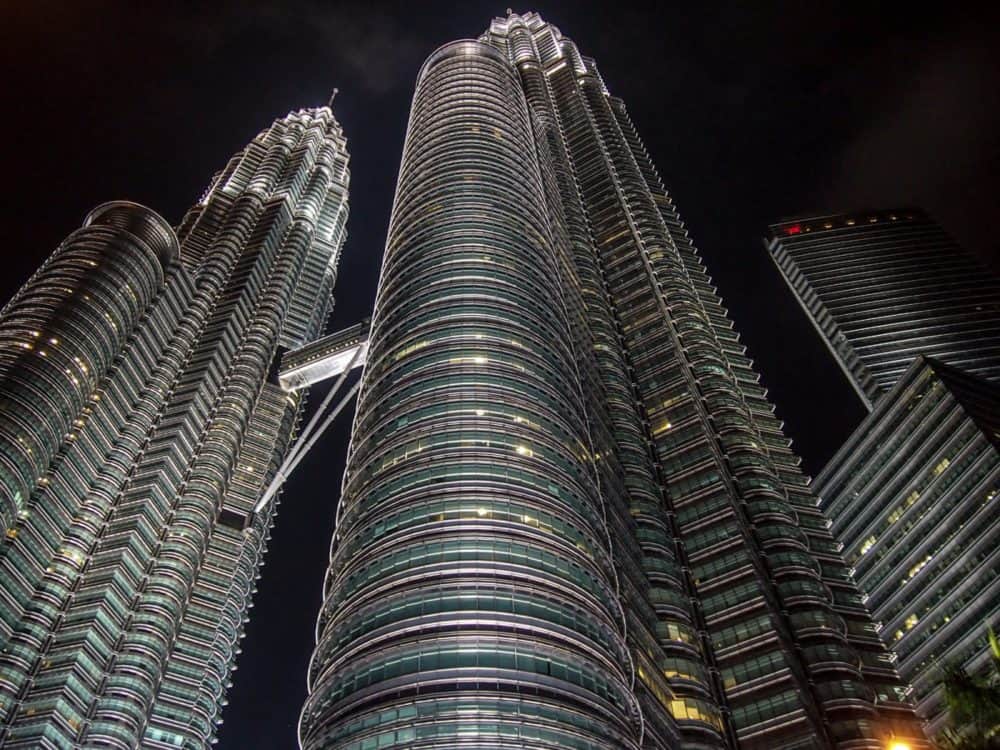 Looking up at a pair of tall skyscrapers with black night sky behind them. The two tours are connected via a covered passageway about halfway up. A few lights are visible in the windows of the building.