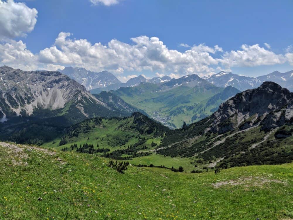 View from the top of a mountain in Liechtenstein, with many other mountains visible in the mid and far distance that have grassy slopes lower down and rocks with small amounts of snow on top.