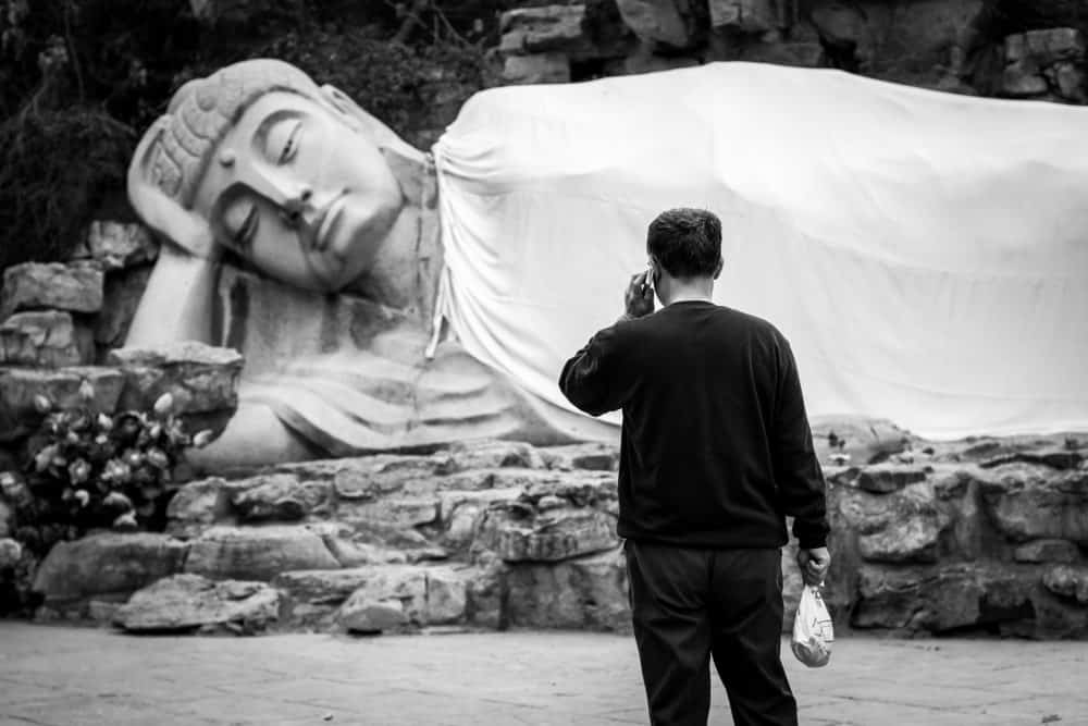 Black and white photo of a man standing in front of a large reclining Buddha statue on a rock platform. Man has a small bag in one hand and is holding a phone to his ear with the other hand.