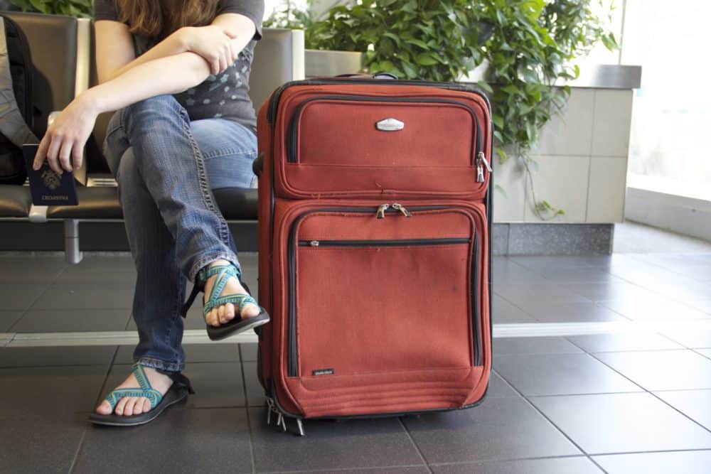 Red suitcase standing vertically alongside a person sitting on a chair with passport in hand.