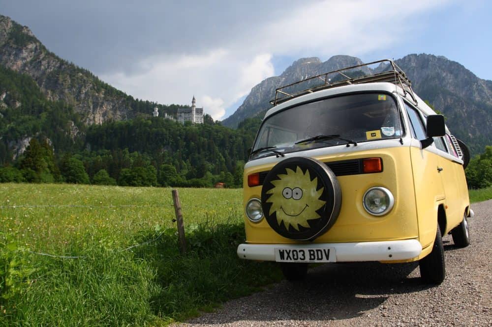 Old yellow campervan with stylized sun on the spare wheel cover on the front, parked on a gravel road beside a field with a castle and mountains in the background