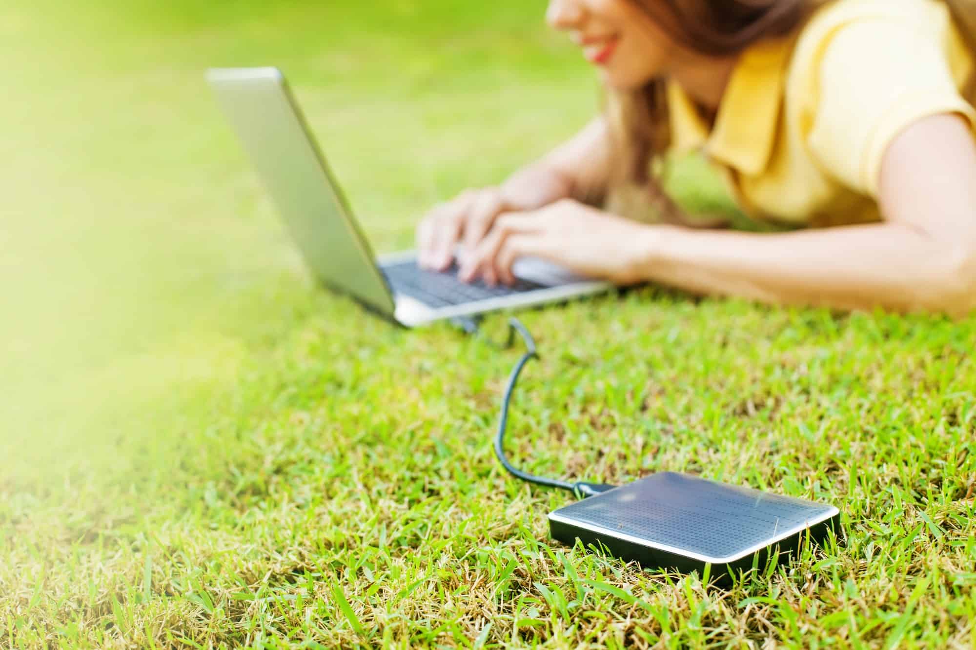 Woman lying on grass using laptop with portable hard drive connected to it