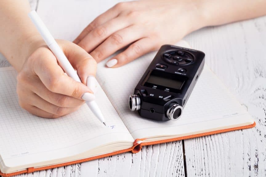 Person writing in notebook with voice recorder sitting alongside