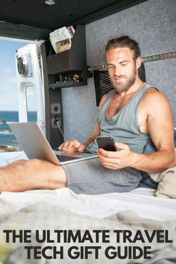 Man inside campervan with laptop and phone, with text "The Ultimate Travel Tech Gift Guide" overlaid at bottom