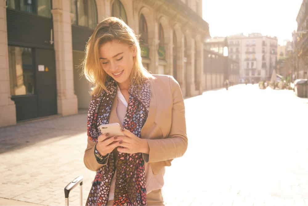 Junge Frau mit Koffer, die Telefon auf Stadtstraße mit Text überprüft "Überprüfung von Airalos eSIM für Reisende" an der Spitze