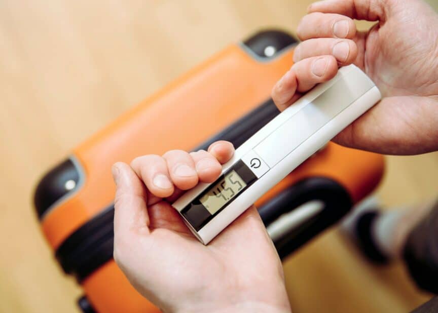 Handheld luggage scale with digital readout above orange suitcase