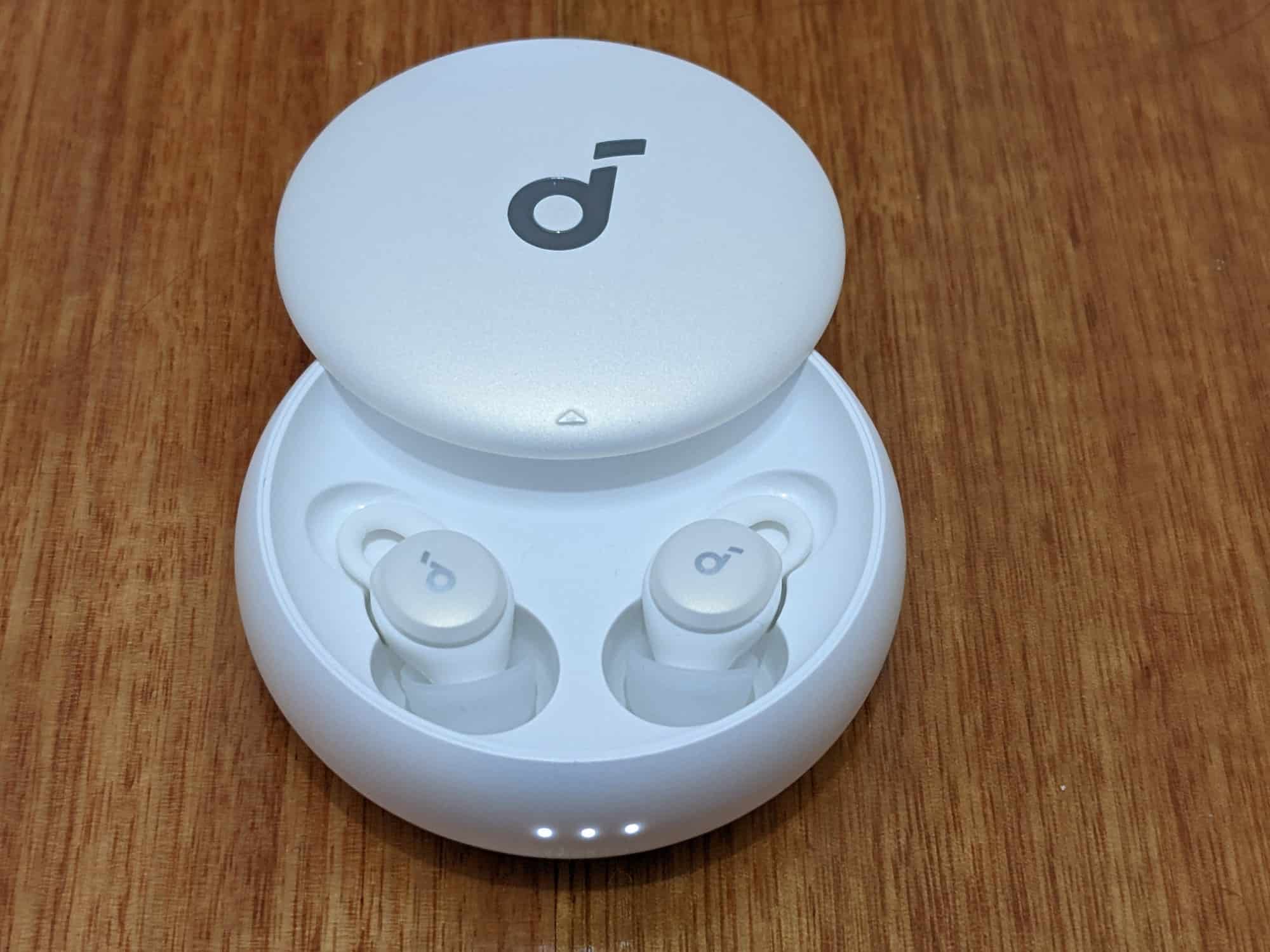 Round white Soundcore Sleep A10 earbuds case on a wooden table, with both earbuds inside the case and the charging lights on the front illuminated