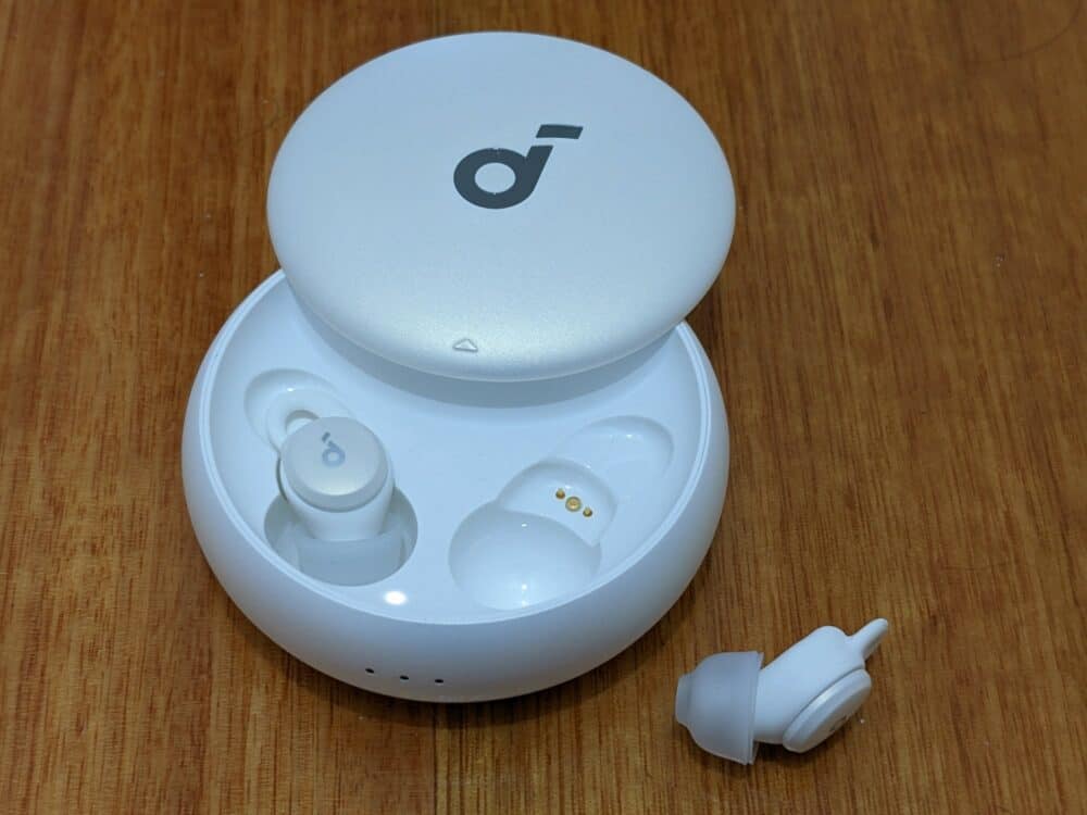 Round white Soundcore Sleep A10 earbuds case on a wooden table, with one earbud inside the case and the other sitting alongside on the right