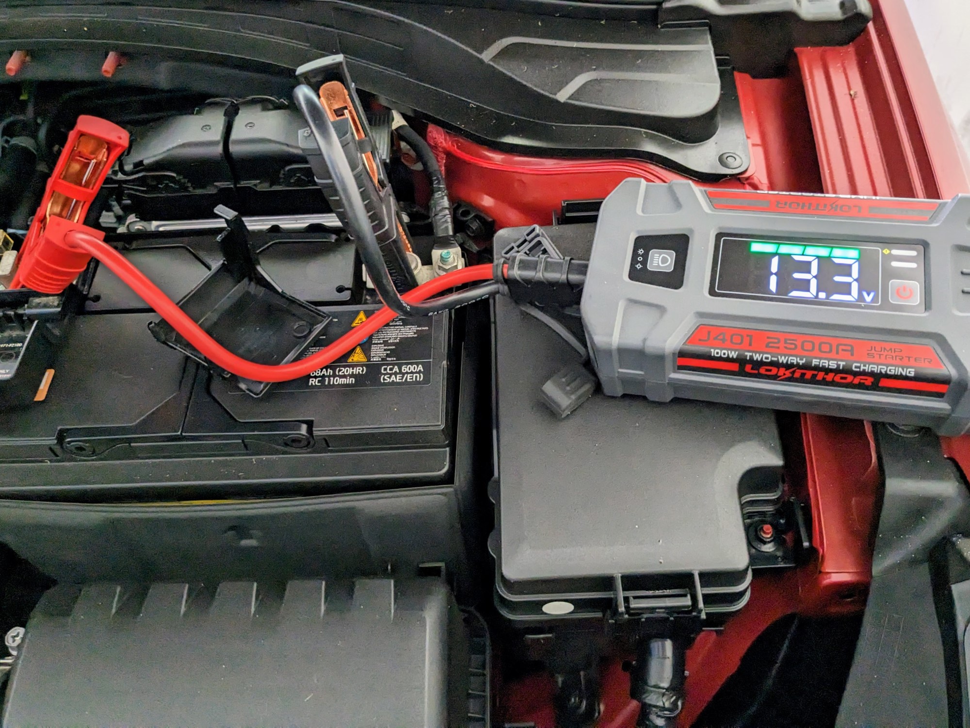 Lokithor J401 portable jump starter with leads connected to car battery. 13.3v measurement visible on display.