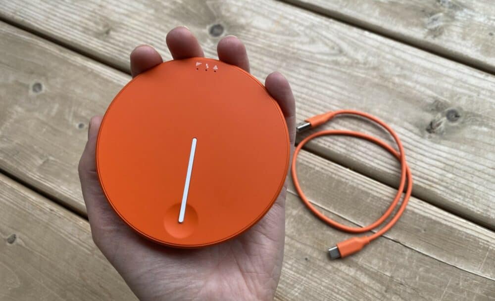 A round, bright orange device with a thin white stripe down the middle and icons around one edge, being held in a hand above a wooden deck with an orange cable lying on it.