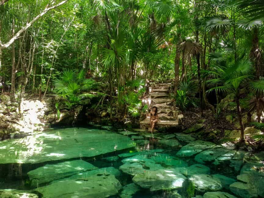 Woman sitting on edge of rough stone steps amidst jungle alongside a cenote in Mexico