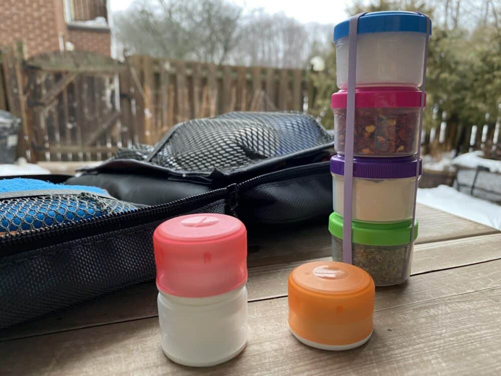 Four round containers filled with spices stacked on top of each other on an outdoor table, with an elastic band around them. Smaller containers in the foreground, with a packing cube in the background.