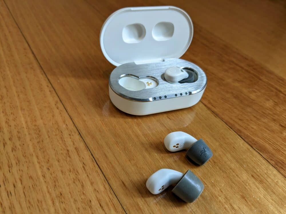 QuietOn 3.0 earbud at front, QuietOn 3.1 in middle, charging case at rear, all on wooden floor.