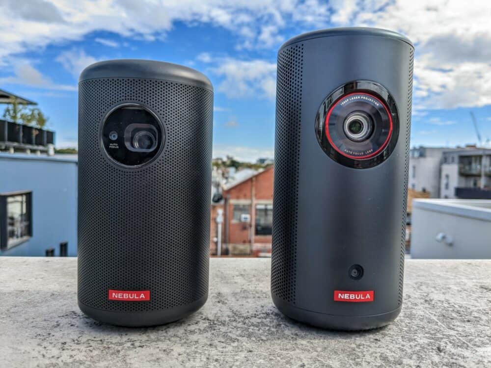 Anker Nebula Capsule 2 and Nebula Capsule 3 portable projectors beside each other on a stone wall with blurred urban landscape in background