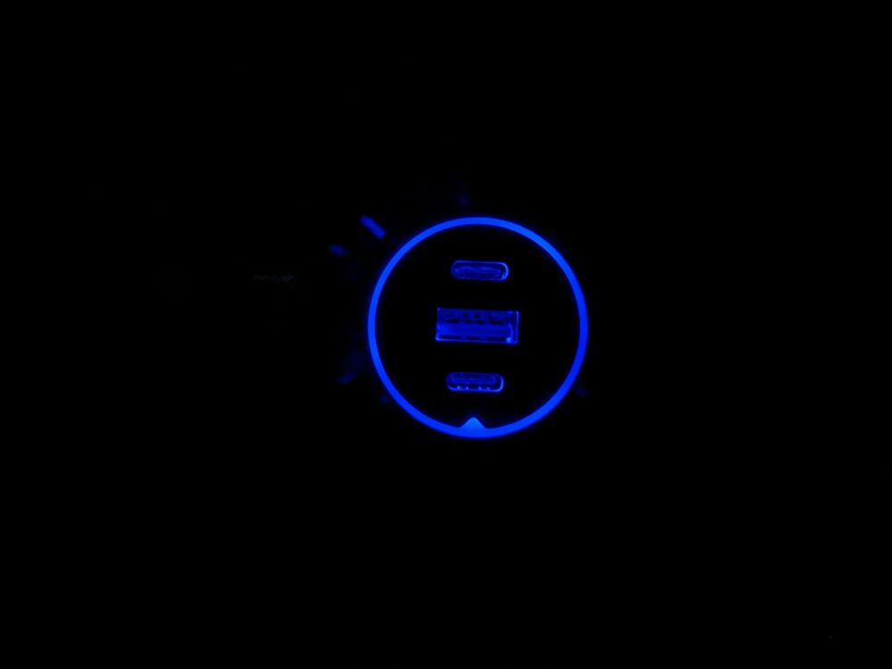 Car charger illuminated in the dark, with a blue ring around the edge and the three USB ports illuminated in blue from inside