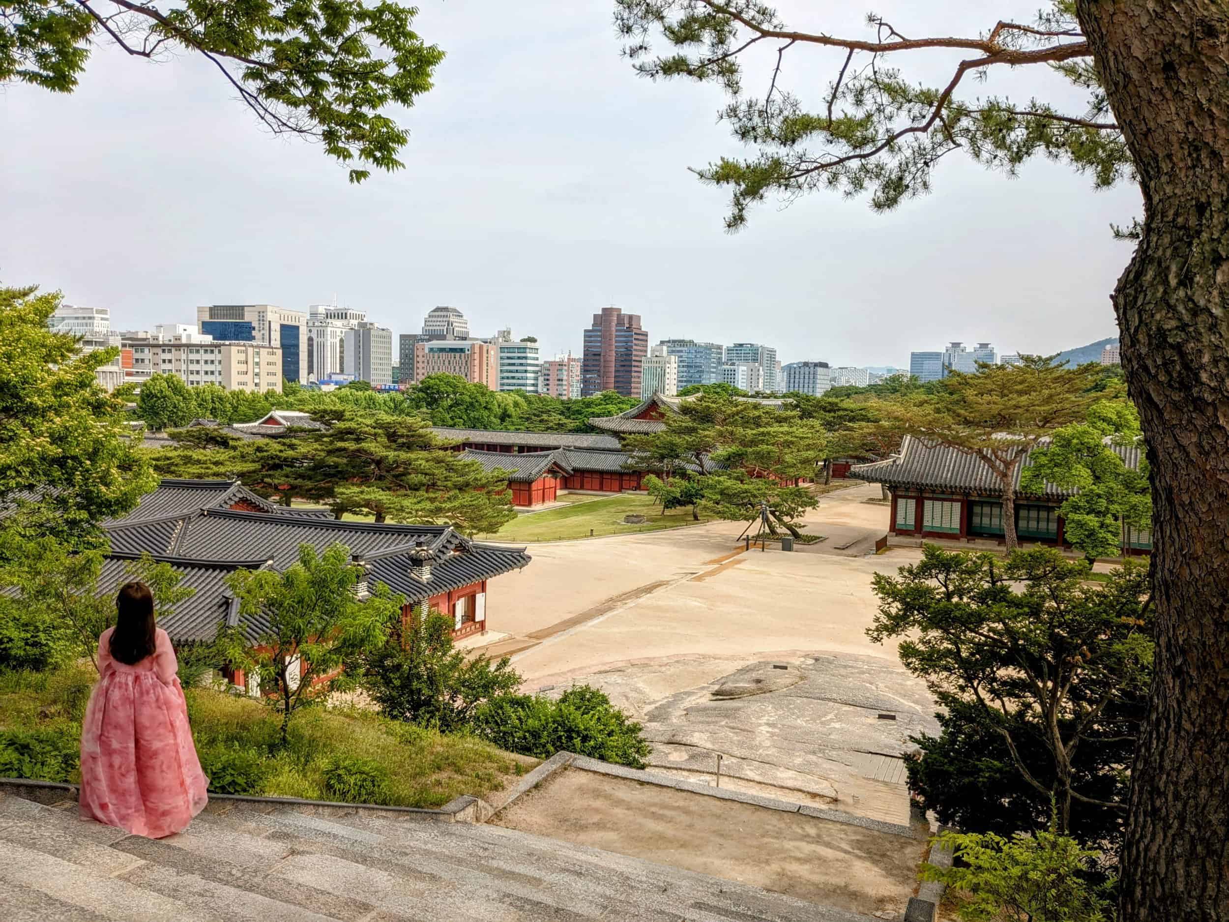 Woman in traditional hanbok dress standing on steps looking out towards buildings of Changgyeonggung Palace, with Seoul cityscape in background