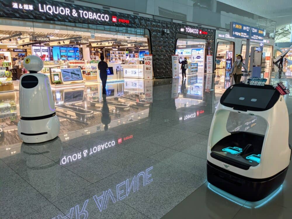 Two robots moving around in a terminal at Incheon Airport in Seoul, South Korea. Retail stores in the background