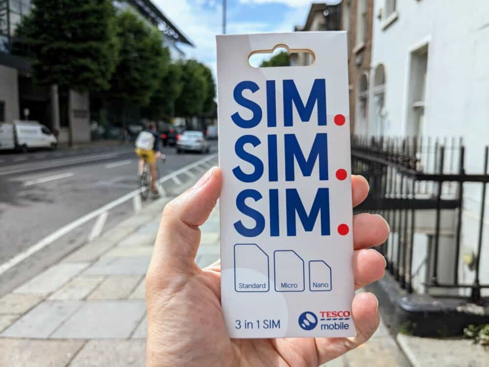 Tesco Mobile SIM card package being held in hand, with blurred city scene in the background.