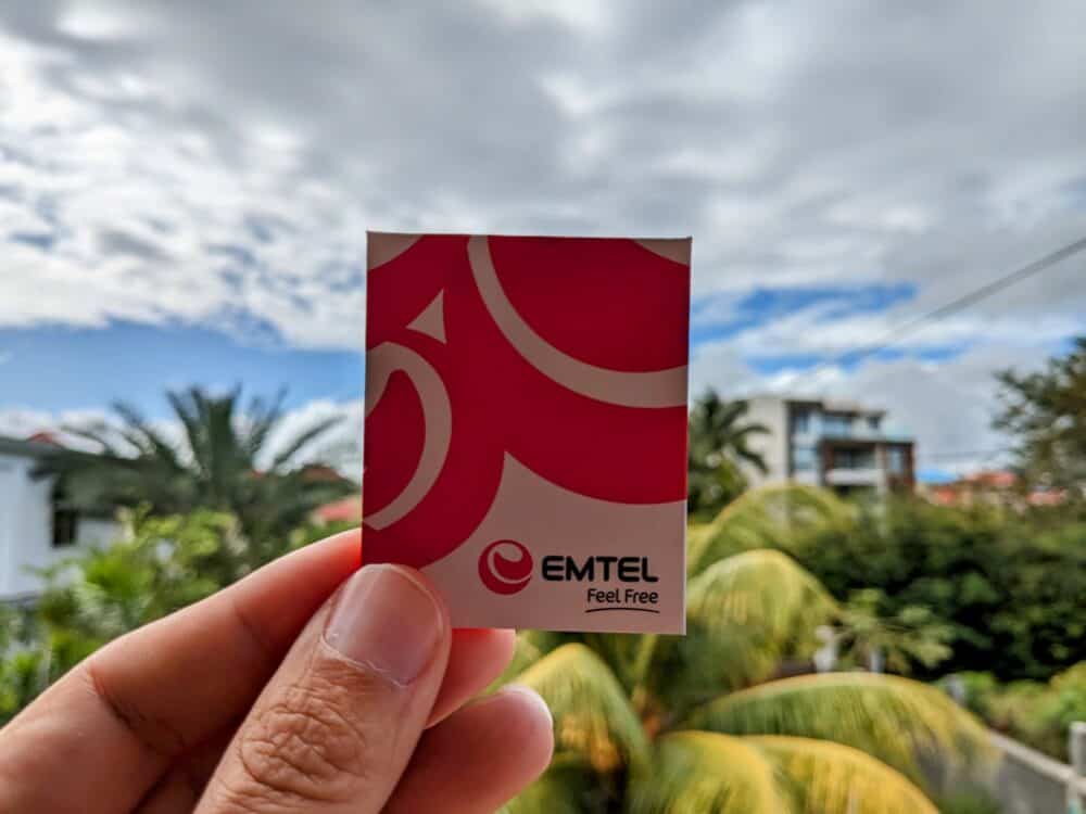 Emtel SIM card package being held in hand with blurred palm trees and buildings in background