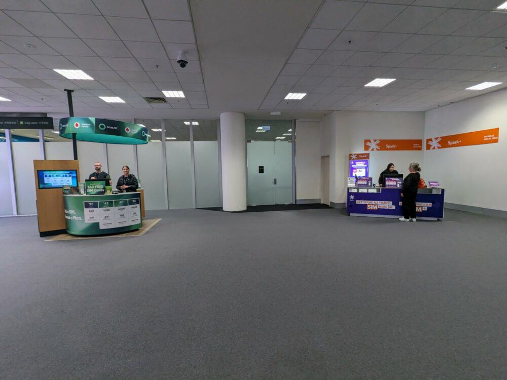 Two small counters in a large, otherwise empty carpeted space at an airport. Counter on left has branding from One NZ and a range of SIM card prices listed, with two staff standing behind. The counter on the left has Spark branding, one staff member, and one customer