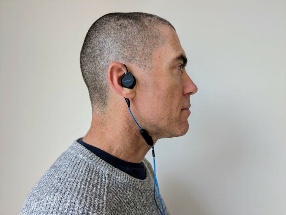 Side view of man in grey sweater and blue t-shirt with a flat earpiece in his right ear. A cable loops around his ear down below his neck. with a black remote control at throat level.