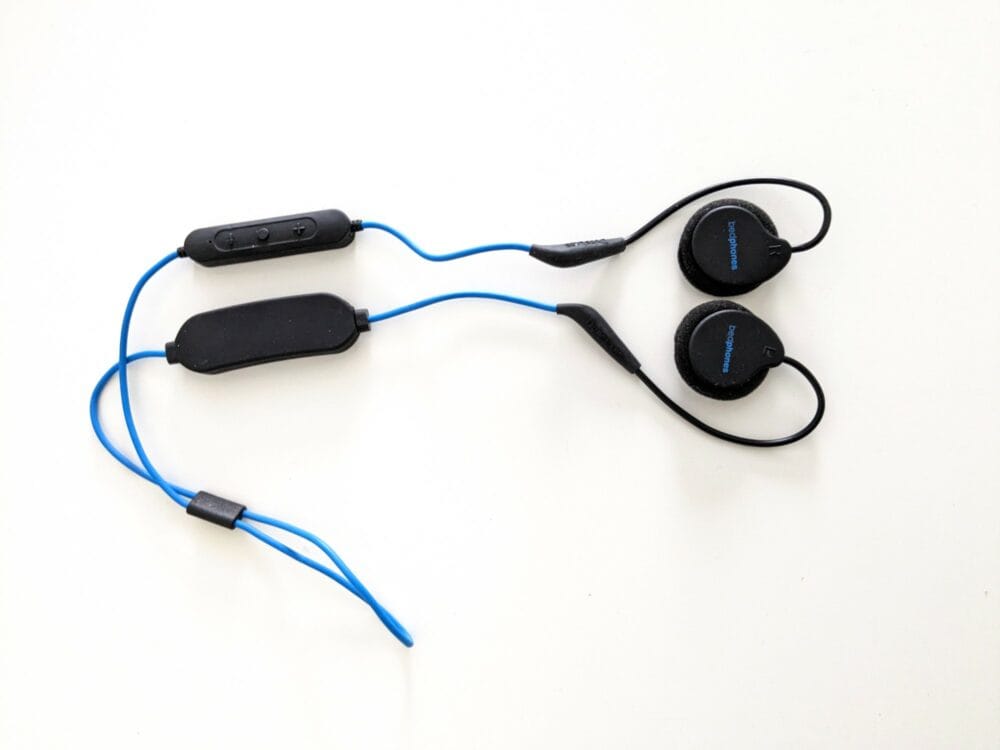 Wireless headphones with flat earpieces and cables that loop around each ear and connect below. Black battery holder on top section of cable, black remote control on bottom section.