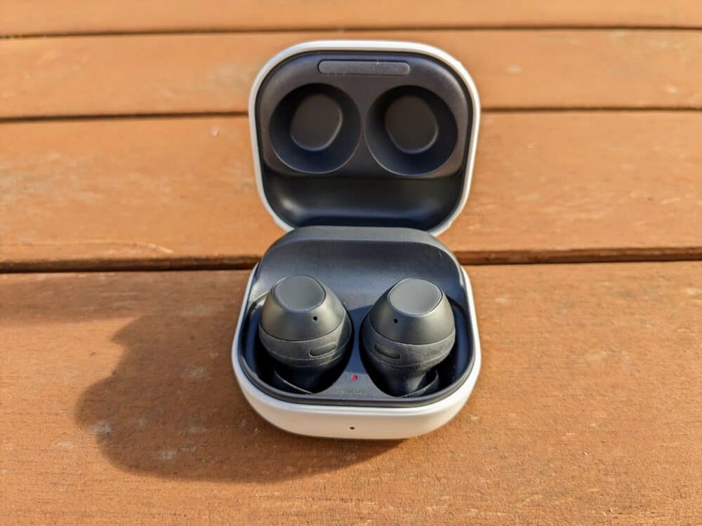 Small square white charging case on a wooden table, with pair of black earbuds inside.