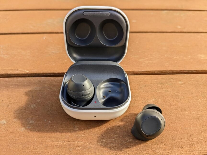 Small square white charging case on a wooden table, with one black earbud inside. Other black earbud sitting alongside.