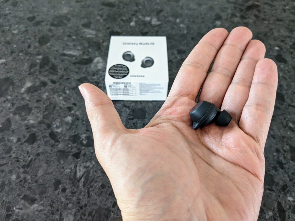 Hand holding single black earbud, with blurred Galaxy Buds FE box visible on a patterned kitchen benchtop behind. 