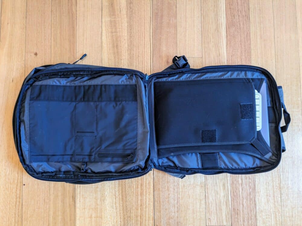 Open daypack on wooden floor with the ends of a keyboard and laptop visible above their storage sections.
