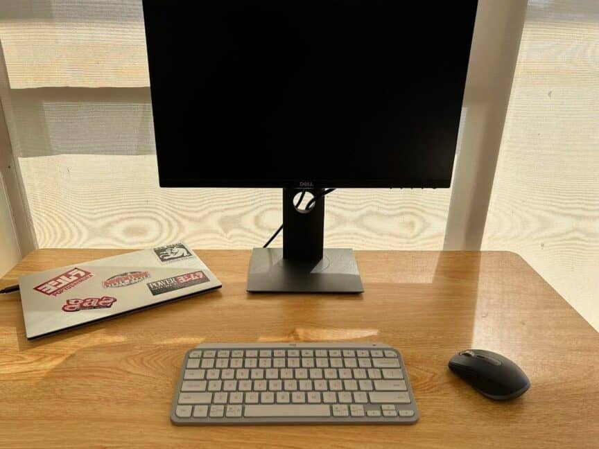 Closed laptop, monitor, keyboard, and mouse on a small wooden desk. Several stickers on top of laptop.
