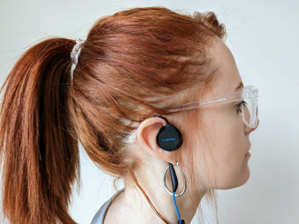 Side view of woman with red hair, glasses, and a hoop earring wearing a flat earpiece in her right ear. A cable loops over her ear and down towards the ground.