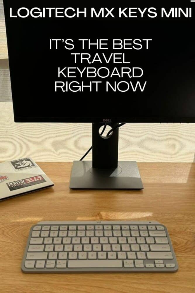 Closed laptop, monitor, keyboard, and mouse on a small wooden desk. Several stickers on top of laptop.. Text "Logitech MX Keys Mini" and "It's the Best Travel Keyboard Right Now" overlaid at top