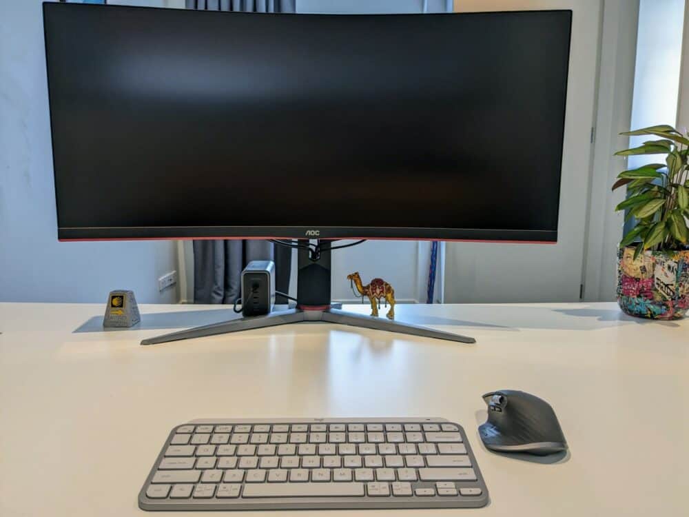 Small keyboard, mouse, and monitor on a white desk in a home office. Potted plant and a few small decorations on the desk.