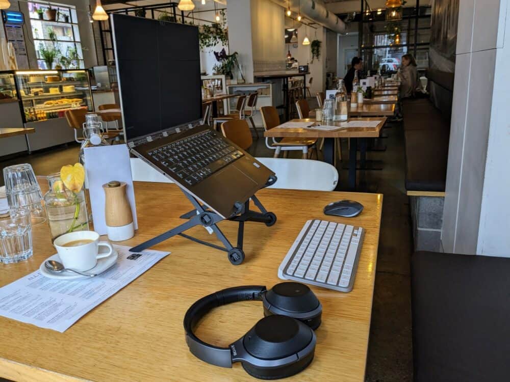 Laptop on a stand, headphones, keyboard, and mouse on a table at a cafe, with coffee cup, menus, and glasses behind. Typical cafe scene behind, with several tables and a few customers. Display case with baked goods and drinks visible to the left.
