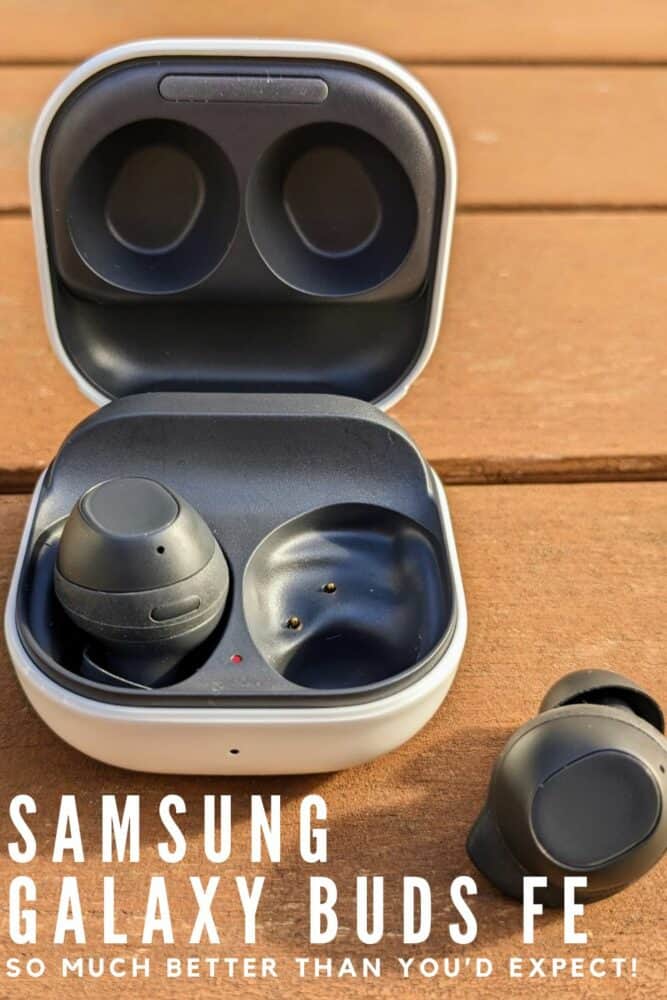 Wireless earbuds case on wooden table, with one earbud in the case and one sitting alongside. Text at bottom reads "Samsung Galaxy Buds FE: So Much Better Than You'd Expect"