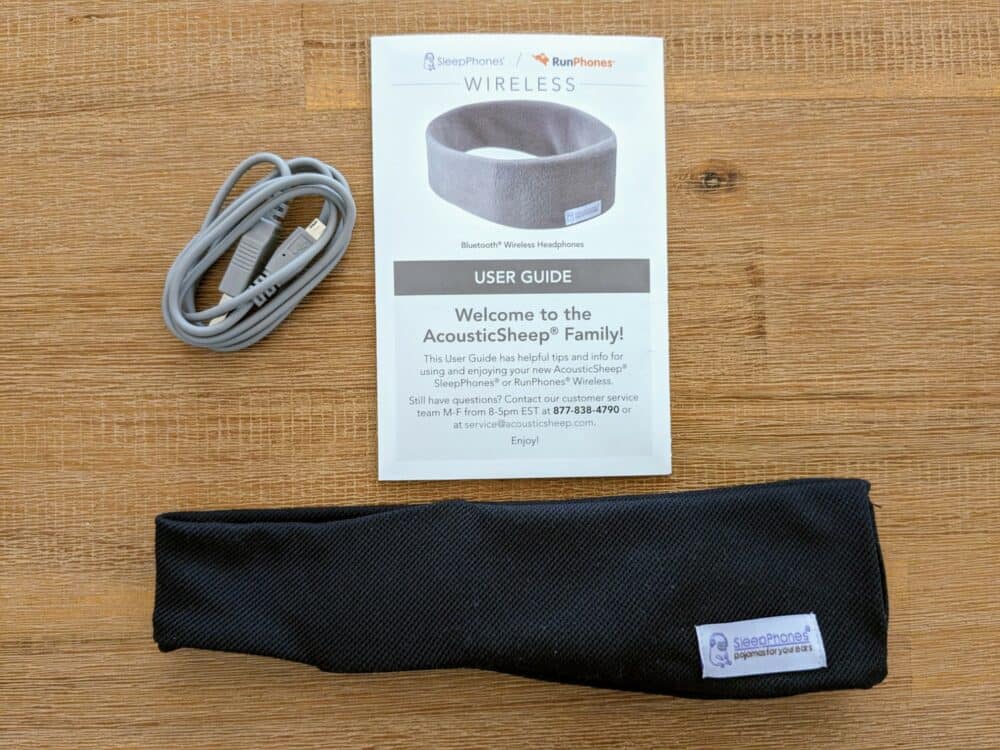 Black headband, grey charging cable, and instruction booklet for SleepPhones Wireless, all sitting on a wooden table. 
