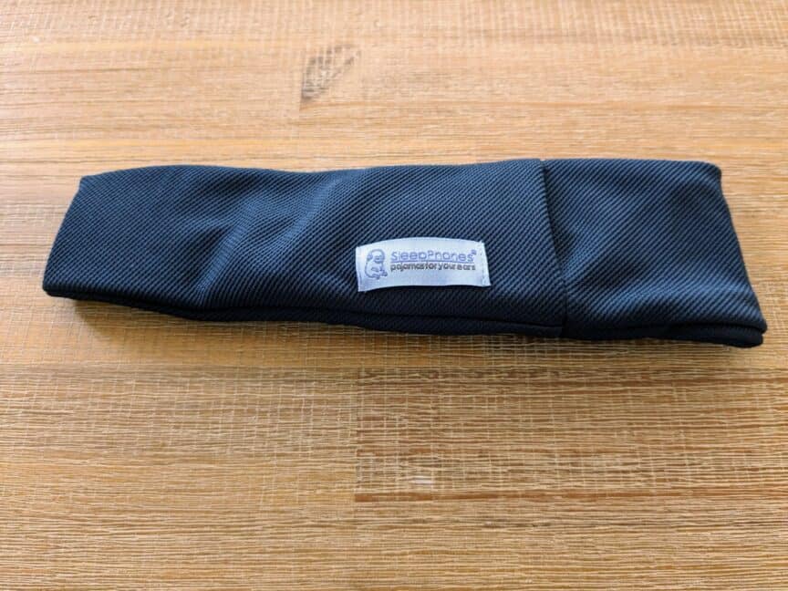 Folded headband made from breathable material lying on a wooden table. Label visible that says "SleepPhones: pajamas for your ears"