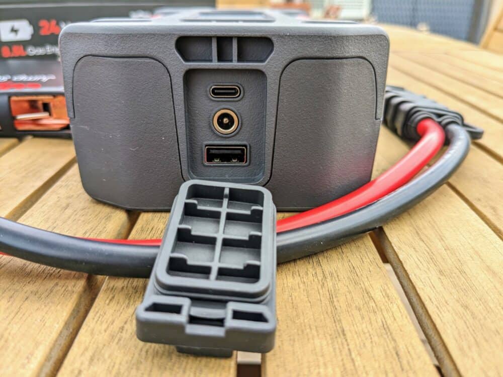 Close-up of USB C, DC, and USB-A sockets on one end of a J401 portable jump starter that is sitting on a wooden table. Jumper cables are looped around the device, with the product box (blurred) visible behind.